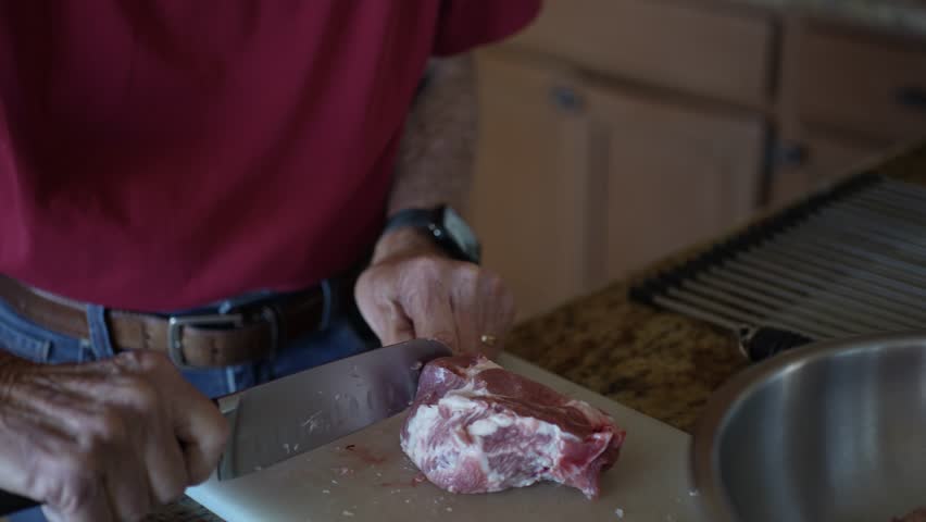 Senior man prepares meat by trimming fat and cutting pieces in his kitchen with a knife. He is focused on the task at hand.