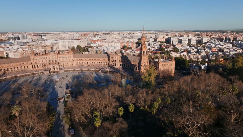 Aerial view of Plaza de Espana in Seville, Spain. Twin towers, central fountain, patterned bridges, and Parque de Maria Luisa trees in warm late afternoon light.