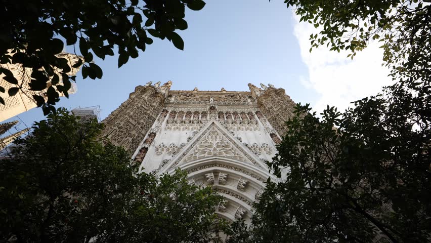 Low angle view of Seville Cathedral portal with tracery, statues, pointed arches, and orange trees. Giralda tower peeks left in late afternoon natural light.