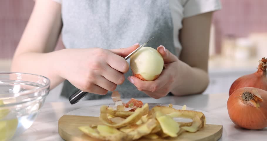 Woman peeling fresh onion at white table indoors, closeup. Camera moving forward