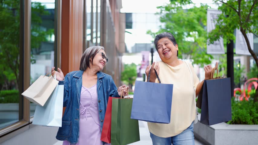 Two happy Asian senior women walking outdoors with shopping bags. Attractive older friends enjoy leisure time together, smiling and carrying purchases after shopping in modern department store or mall