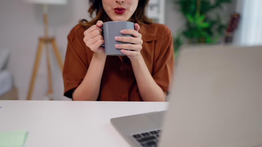 Close up of female freelancer drinking coffee while working from home. Attractive young businesswoman or student sitting on table using laptop device learning online in cozy living room in house.
