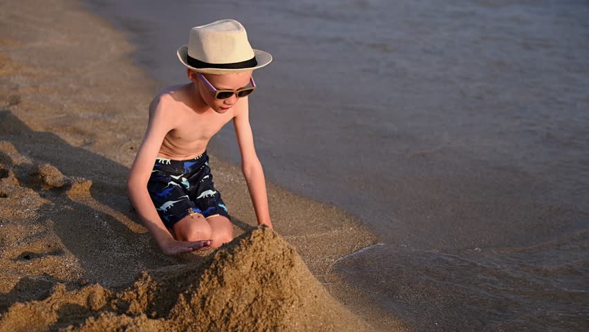 Boy on sandy beach looks out at sea while creating sand sculpture. Young child enjoys relaxing summer vacation. Concepts of children