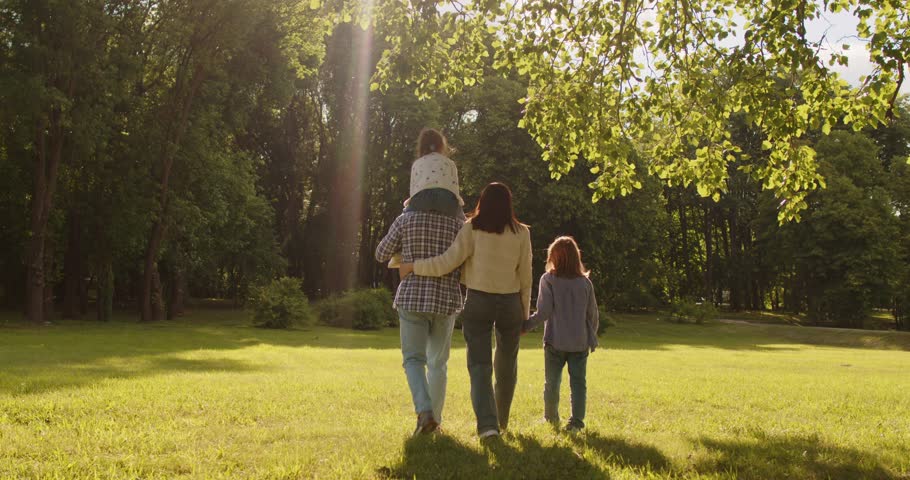 Back view of lovely family walking slowly on green grass, admiring beautiful nature and sunshine. Father giving piggy back to small daughter. Outdoor video of parents and two children in park