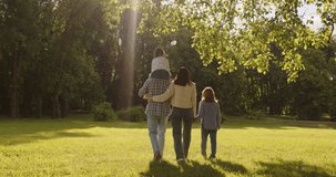 Back view of lovely family walking slowly on green grass, admiring beautiful nature and sunshine. Father giving piggy back to small daughter. Outdoor video of parents and two children in park - Powered by Shutterstock - Get 15% off with code: PIKWIZARD15