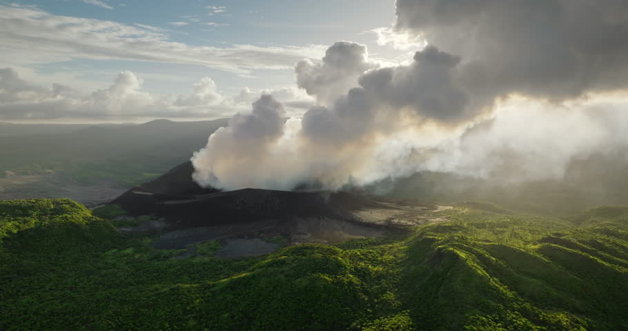 Mount Yasur volcano actively erupting in Vanuatu, releasing thick white smoke and ash into the sky, surrounded by lush green tropical vegetation on Tanna island. Aerial view drone flight panorama