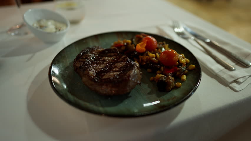 Grilled steak served with vegetables and cherry tomatoes on elegant plate in restaurant setting. Gourmet dinner meal prepared for fine dining experience on white tablecloth.