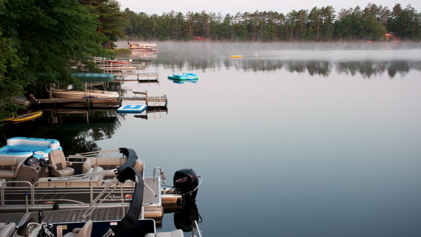 Morning mist drifts over a calm lakeside with boats and docks reflecting on still water.