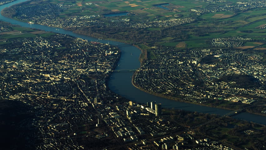 Aerial view of Rhine River flowing through urban landscape near Cologne in North Rhine Westphalia, showing bridges, residential districts and countryside fields surrounding major city.