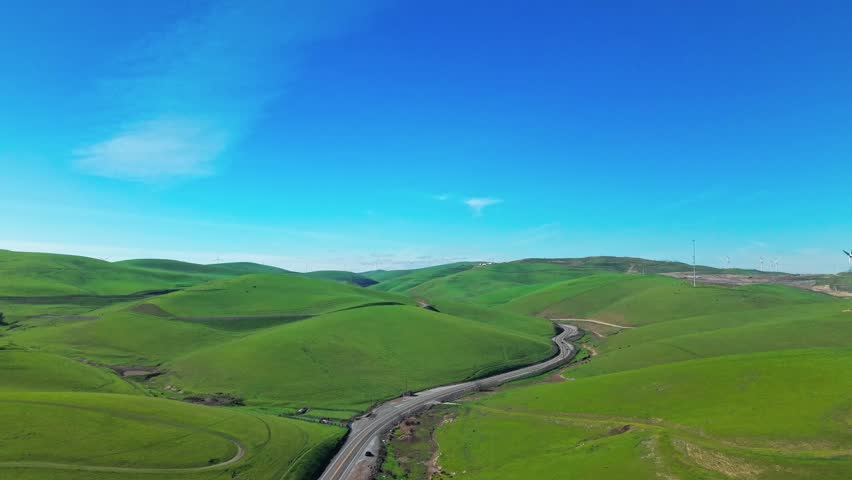 Aerial drone footage flying over the vibrant green rolling hills of Altamont Pass, California, revealing a winding road and distant wind turbines under a clear blue sky on a sunny day