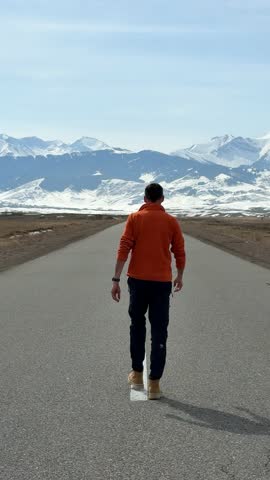 Rear view of a man walking on a deserted road towards snowy mountains, spreading his arms in a gesture of freedom and joy