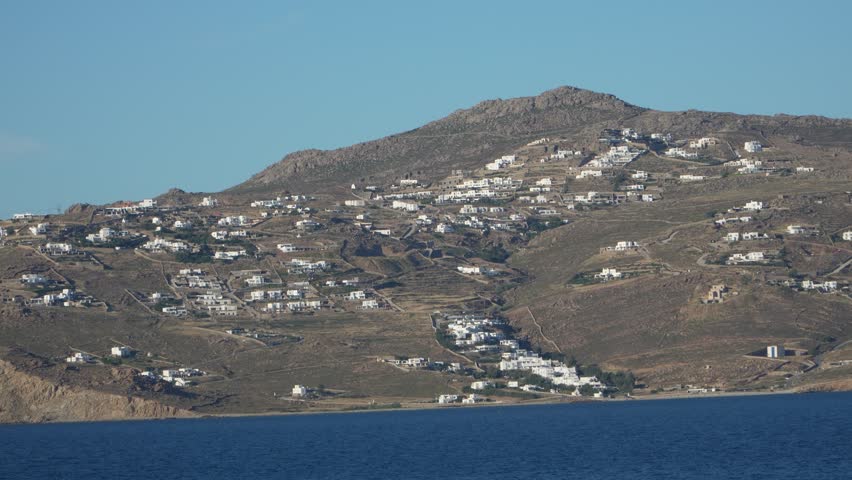 Traditional white Cycladic houses scattered across brown hillside overlooking blue Aegean Sea in Mykonos, Greece. Scenic coastal landscape view from departing cruise ship.