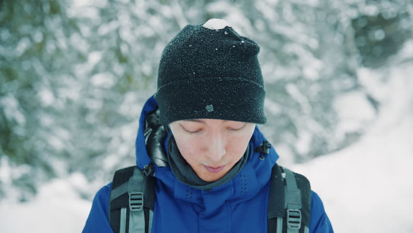 Close up portrait of an asian man raising his head and smiling at the camera during a cold winter hike. The adventurer is exploring a beautiful snowy forest during a snowfall