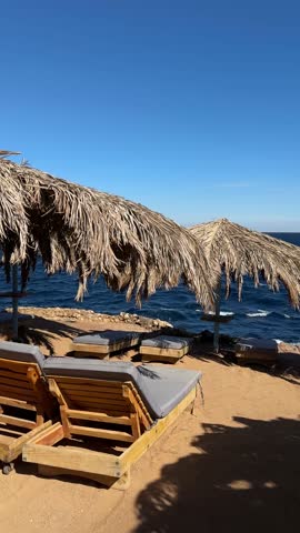 Vertical scenic view of wooden loungers and straw beach umbrellas beside rocky shoreline with blue Aegean Sea creating relaxed summer resort atmosphere in Greece