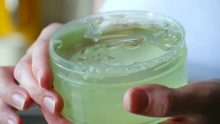 Female hands holding jar of transparent green aloe vera gel, scooping out product with finger for moisturizing skincare routine, showing smooth, cool texture of cosmetic. Use jar with aloe vera gel