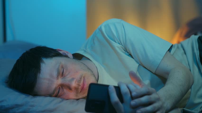 Young man lying in bed late at night, scrolling smartphone, feeling sleepy, eventually turning off device, go to sleep in dark bedroom with blue, warm light. Man using phone at night before sleeping