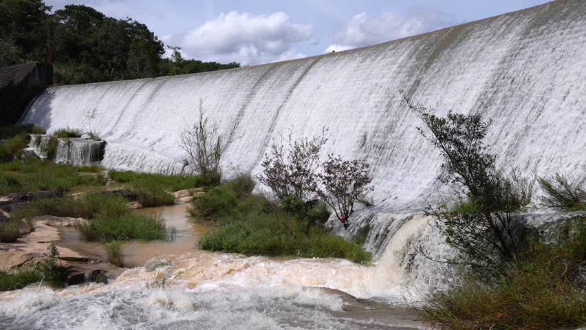 A wide, concrete dam overflows with rushing white water, cascading into a rocky, vegetation-filled riverbed under a cloudy, bright sky.