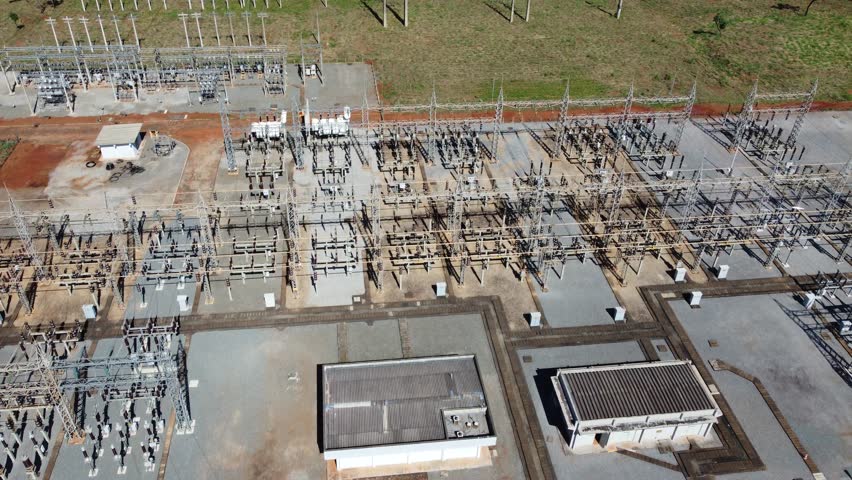 Aerial tracking shot passing over a transformer station with a slow pan up to reveal a suburban landscape and green fields in Goiania, Brazil.