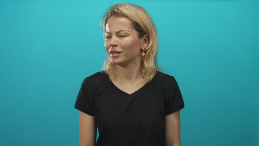 Young blonde woman holding lower back and wincing at pain against turquoise studio wall; discomfort.
