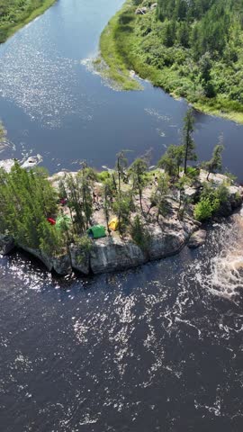 Old Woman Falls island campsite on the Manigotagan River in Manitoba surrounded by rapids. Vertical aerial view backs away from the island camp revealing tents, canoes and the river.