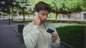 Young hispanic man holding phone to ear while inspecting a credit card on a street with trees and walkway; financial worry fraud. - Powered by Shutterstock - Get 15% off with code: PIKWIZARD15