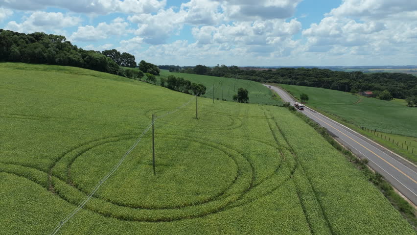 Rural farmland landscape with highway traffic, green crop fields and preserved woodland under a cloudy blue sky.