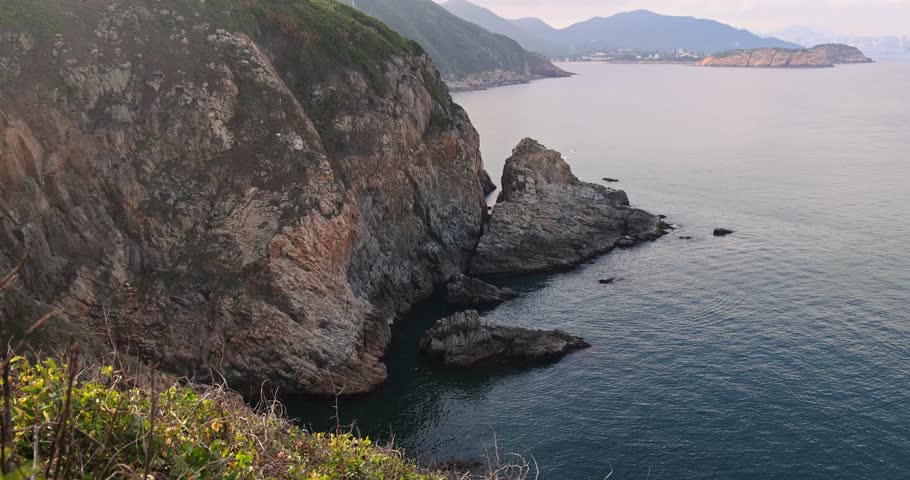 Scenic steep rocky cliffs meeting calm ocean. Distant mountains under soft hazy light. Natural coastal landscape.