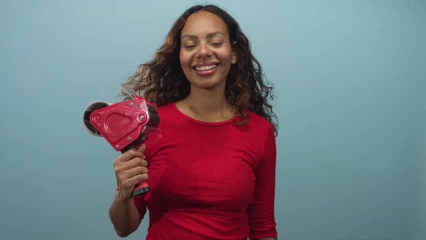 Young african american woman holding red packing tape gun and making i love you hand gesture in blue studio; playful joy.