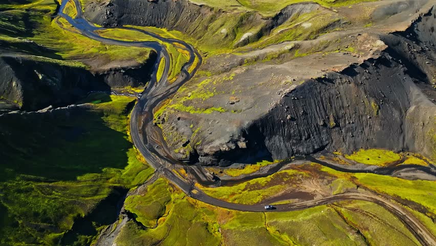 Drone panorama of a tiny car driving a winding highland road through Iceland’s green summer valley and dark volcanic cliffs. Remote living, freedom, and adventure travel concept.