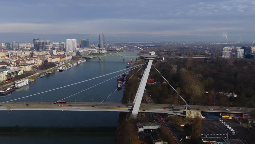Aerial view in Bratislava shows Most SNP UFO bridge, Old Town, St Martins Cathedral, glass towers, green truss bridge, Apollo Bridge, buses, cars, boats, overcast light.