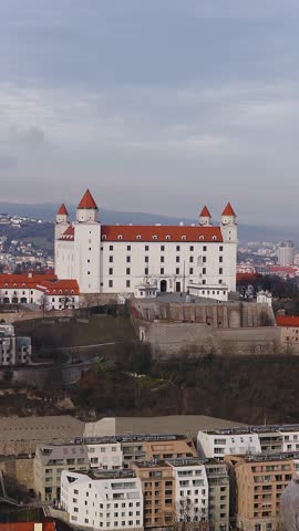 Aerial drone pan shows Bratislava Castle with four towers and orange roofs, stone bastions and ramparts, modern apartments, and city skyline in soft overcast light.
