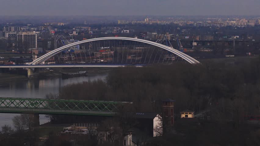 Aerial telephoto view of the Danube in Bratislava. Apollo Bridge rises behind the Old Bridge. Boats and vehicles move under hazy late afternoon light.