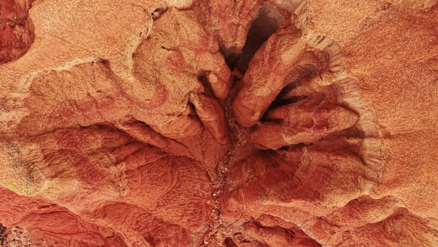 A canyon features various red rock formations with unique shapes and lines under bright sunlight. The texture of the rocks is visible, showing the Earth