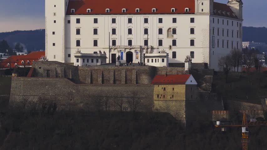 Aerial pan shows Bratislava Castle with four corner towers, red roof, stone bastions, terrace visitors, a crane, and a distant tower in late afternoon light.