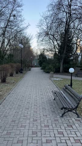 First person walking shot along a park path while looking around the environment. The camera turns from green lawns and trees to residential buildings visible in the distance under bright blue sky.
