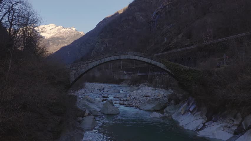 Aerial view shows a single span stone arch bridge over a turquoise alpine river in Italy, with arched retaining walls, wooded slopes, and a snow capped peak.