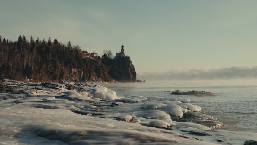 Split Rock Lighthouse rises above frozen Lake Superior shoreline at sunset with dramatic ice formations sea smoke and misty winter atmosphere in Minnesota - ground level view