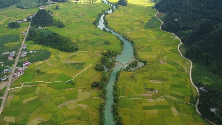 Aerial drone reveal of winding river flowing through patchwork rice fields with rural roads, small villages and green valley landscape surrounded by limestone hills in Ninh Binh Vietnam