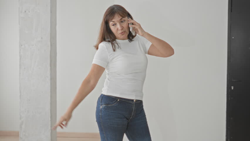 Woman in white t shirt and jeans on smartphone, hand to ear for call, standing by concrete column in building interior; calm domestic moment.