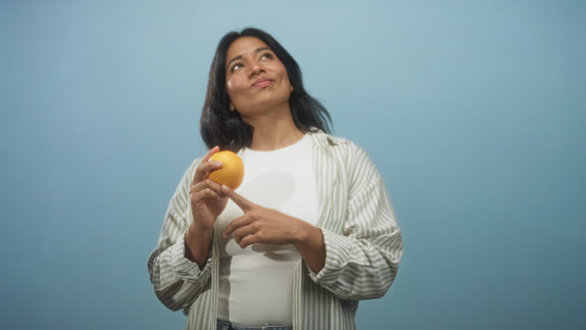 Woman holding an orange in her left hand and pointing her right index finger to lips while wearing a white top and striped overshirt against a blue studio wall; curiosity reflection.
