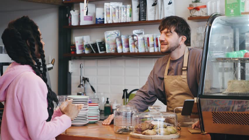 A barista interacts with a customer at a cafe, exchanging smiles while serving coffee and pastries during a busy morning.