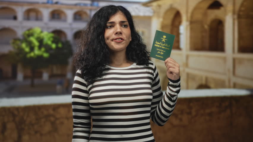 Young woman holds passport and presses hand to forehead in building in an old town square; frustration uncertainty.