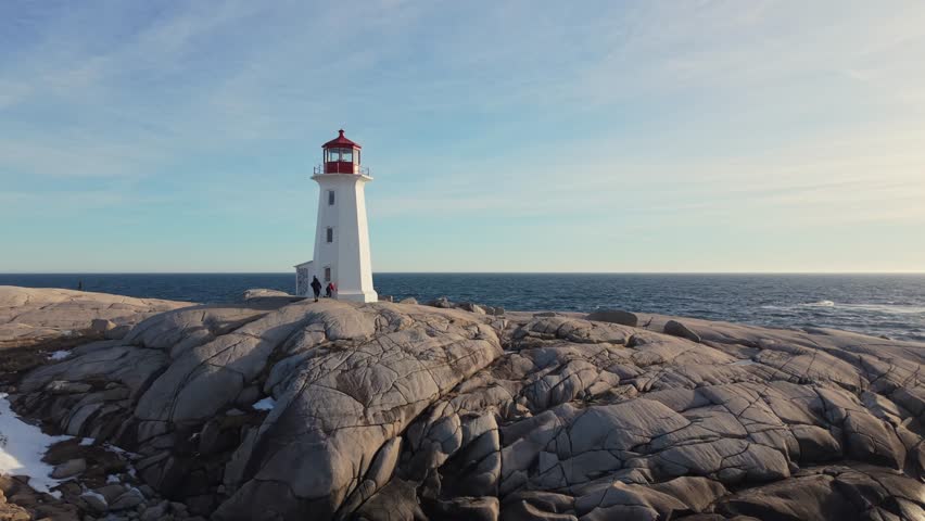 Drone aerial shot: Peggy's Cove lighthouse and snowy granite rocks at sunset over Atlantic Ocean. Halifax suburb Nova Scotia Canada, dramatic winter seascape with golden light 4K.