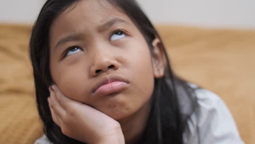 Upset little Asian girl lying on stomach on bed, chin resting on hands with a sulking face, unhappy child feeling grumpy or bored in bedroom