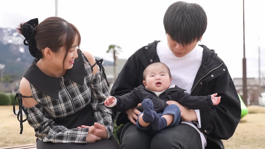 Man holds baby gently in park setting. Child smiles softly, eyes wide with wonder. Bright yellow tent stands behind bare trees. Distant pine tree adds depth to natural backdrop. Sunlight warms scene