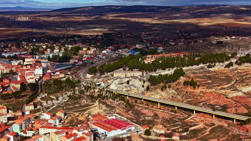 The town shows houses and buildings scattered along the valley. Roads connect different areas. Hills and open land are visible in the background under a clear sky.