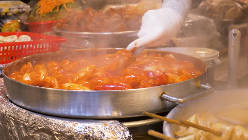 Slow motion close-up of person wearing gloves using spoon utensil mixing tteokbokki fish cake soup served within sauce broth in pan at Gwangjang night street markets in Seoul city South Korea cuisine