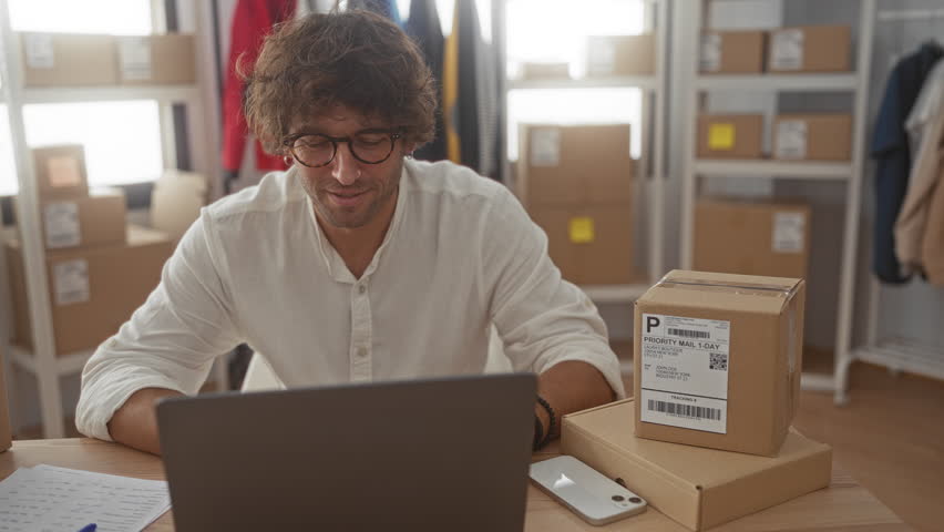 Man working on laptop at desk surrounded by cardboard shipping boxes and labeled parcels in a packing building; entrepreneurship focused.