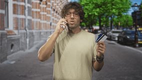 Man holding creditcard in hand and talking on phone on street outside brick building; concern identity theft. - Powered by Shutterstock - Get 15% off with code: PIKWIZARD15