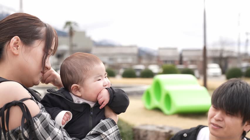 Man holds baby gently in park setting. Child smiles softly, eyes wide with wonder. Bright yellow tent stands behind bare trees. Distant pine tree adds depth to natural backdrop. Sunlight warms scene